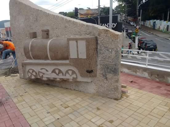 Painel decorativo esculpido em pedra com relevo de locomotiva, instalado em praça pública